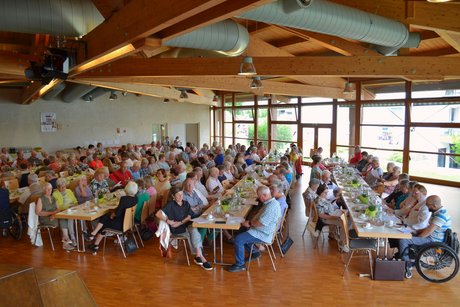 Panorama eines gut befüllten Saals, in dem Menschen an Tischen sitzen und mitsingen.