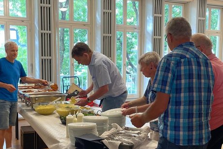 Buffettisch mit Tellern, Besteck, Schalen mit Kartoffelsalat und Warmhalteboxen. Dahinter gibt ein Mann Essen aus, w&auml;hrend davor vier Personen mit Tellern in der Hand warten.