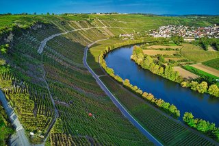 Auf dem Bild ist das Neckartal zusehen, auf der linken Seite sind im Schatten liegende Weinberge und rechts vom Neckar Felder und Weisen in der Sonne.