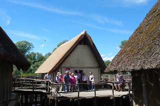 Eine Gruppe Personen steht bei blauem Himmel vor einer Hütte der Pfahlbauten in Unteruhldingen. Die Hütte steht auf Holzpfählen im Wasser am Ufer des Bodensees.