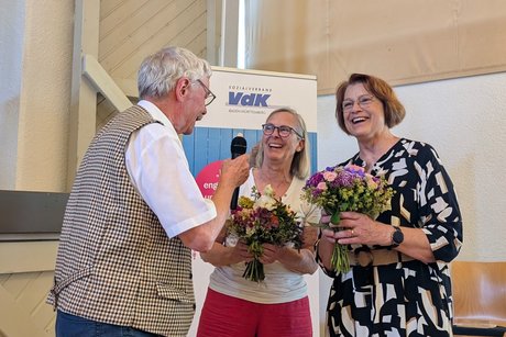Ein Mann im weißen Kurzarmhemd überreicht einer Frau in weißer Bluse einen Blumenstrauß. Daneben steht eine Frau in schwarzem Kleid mit Blumenmuster, die bereits einen Blumenstrauß in den Händen hält. Alle lachen herzlich.