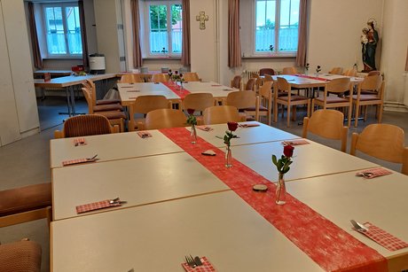 In Gruppen angeordnete Tische, eingedeckt mit rotem Tischband, einzelnen roten Rosen in kleinen Flaschen, Holzscheiben zur Deko, Besteck in rot-weiß karierten Bestecktaschen.