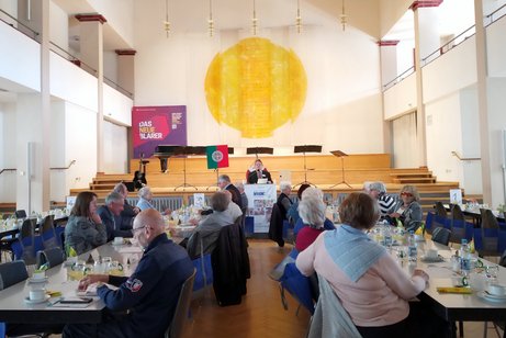 Panoramaaufnahme des Festsaals mit mehreren Tischreihen, an denen zahlreiche Personen sitzen; im Hintergrund steht ein Mann am Rednerpult.