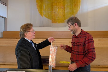 Ein Herr im Anzug &uuml;berreicht einem Mann im roten Hemd eine Wein-Geschenktasche, w&auml;hrend der Mann im roten Hemd herzlich l&auml;chelt.