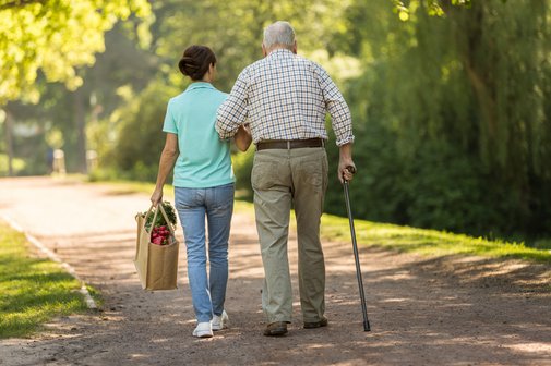 Sozialverband VdK Pflege Beratung Älterer Mann geht an einem Stock und stützt sich ab, eine Pflegerin oder junge Frau geht neben ihm her, sie trägt eine Einkaufstasche mit einer Hand und stützt den älteren Mann mit dem anderen Arm.