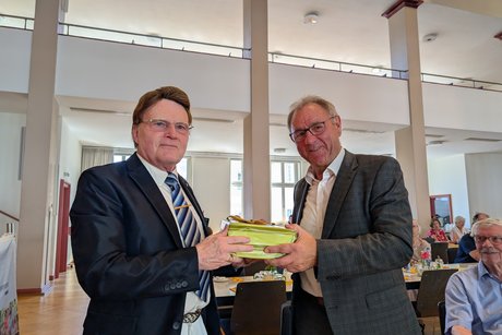 Ein Mann im Anzug mit Krawatte überreicht einem Mann im legeren Anzug ohne Krawatte eine Schachtel.