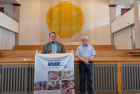 Mann mit blauem Hemd und grauem Jacket am Rednerpult, daneben steht ein Mann im kurzärmeligen Hemd und schaut ihn von der Seite aus an.