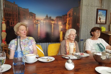 Drei Frauen sitzen vor leeren Tellern und Kaffeetassen und blicken zu ihrem Gesprächspartner gegenüber; an der Wand hängt ein Leinwandfoto eines Kanals mit historischen Gebäuden, vermutlich in Venedig