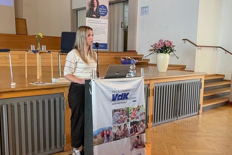 Nicht ganz eindeutig. Besser: Junge Frau mit einer wei&szlig;en Bluse, die mit vier d&uuml;nnen dunklen Querstreifen versehen ist, steht an einem Rednerpult mit VdK-Banner und spricht in ein Mikrofon.