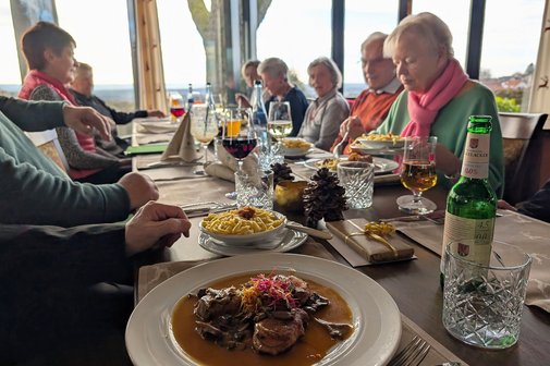 Tisch, an dem mehrere Personen sitzen. Im Vordergrund ein Teller mit Fleisch und Soße, daneben ein kleiner Teller mit Spätzle. Im Hintergrund etwa zehn Personen, leicht verschwommen, die sich unterhalten.
