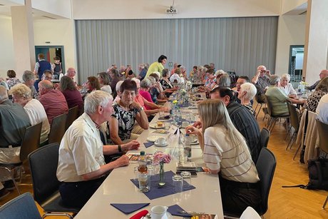 Panorama des Festsaals mit drei Tischreihen, an denen eine bunt gemischte, fr&ouml;hliche Runde von Menschen sitzt und sich angeregt unterh&auml;lt.