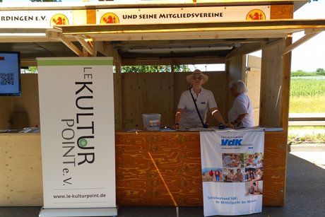 Holzstand mit VdK-Stoffbanner, davor stehen zwei Frauen im sommerlichen Look, eine trägt einen Strohhut.
