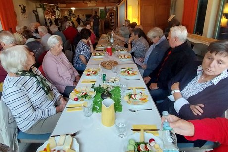 Sommerausflug VdK OV RSKN-Esslingen 2025 Langer Tisch mit mehreren älteren Menschen, vor jedem Teller mit Käsekostproben und Brotkörben in der Mitte, während sie sich unterhalten.