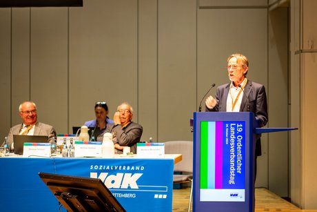 Bernhard Gschwender spricht am Rednerpult, daneben sitzen drei Personen und hören zu