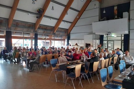 Panorama des Festsaals mit mehreren Tischreihen, an denen viele Leute sitzen.