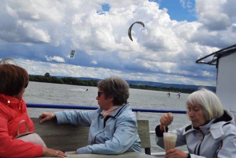 Zwei Frauen auf dem offenen Oberdeck beobachten Kitesurfer auf dem See, daneben sitzt eine Frau am Tisch und trinkt einen Latte Macchiato.