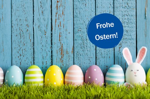 Vor einem h&ouml;lzernen Lattenzaun sind nebeneinander aufgereiht auf einem gr&uuml;nen Rasen viele bunte Ostereier, dazwischen ein wei&szlig;es H&uuml;hnerei mit angeklebten Hasenohren und einem H&auml;schengesicht. Auf einem kreisrunden blauen St&ouml;rer ist die Aufschrift "Frohe Ostern!" zu lesen.