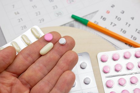 Ausgestreckte Hand mit drei Tabletten in unterschiedlicher Form und Farbe darauf, unter den Hand ist ein Tisch zu sehen auf dem wietere Tablettenpackungen liegen sowie ein Kalender und ein Stift.
