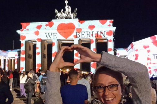 Das Brandenburger Tor bei Dunkelheit mit einer Lichtaufschrift "We Love Berlin", davor die lachende stellvertretende Vorsitzende, die mit hoch erhobenen Armen mit beiden Händen ein Herz formt.