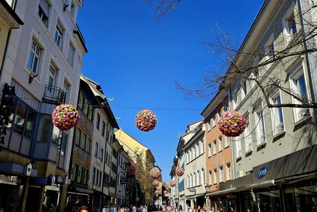 In der Fu&szlig;g&auml;ngerzone in Konstanz sind einige Passanten, hoch oben h&auml;ngen &uuml;berall gro&szlig;e Bl&uuml;tenkugeln gespannt an Seilen.