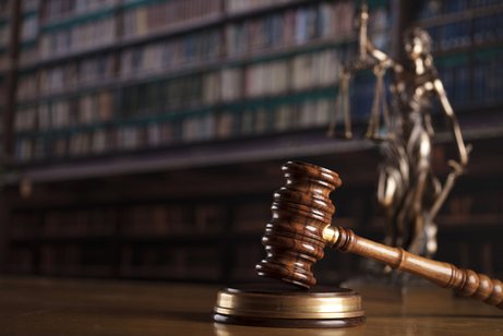 Nahaufnahme eines Richterhammers, dahinter ist eine Figur von Justizia zu sehen, hinter ihr ein B&uuml;cherregal.