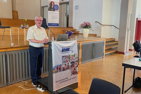 &Auml;lterer Herr mit wei&szlig;em Hemd, kurzen grauen Haaren und Brille steht an einem Rednerpult mit VdK-Banner und spricht in ein Mikrofon.