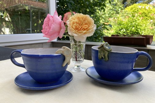 Zwei Kaffeetassen auf einem Tisch, dazwischen eine kleine Blumenvase mit Rosen.