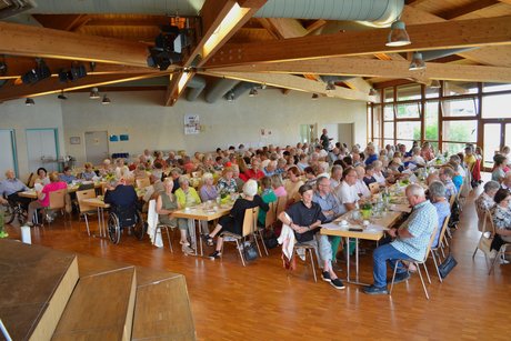 Panorama eines gut befüllten Saals, in dem Menschen an Tischen sitzen und mitsingen.