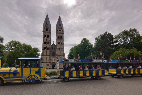 Gelbblaue Lokomotive mit Wagons des Altstadtexpress Koblenz steht vor dem Kastordom , eine Kirche mit zwei Kirchtürmen mit Spitzdach. Personen sitzen in den Wagons. Der Himmel ist bewölkt.