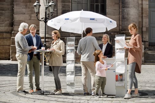 Ein VdK-Infostand in einer Fu&szlig;g&auml;ngerzone, eine Frau spricht mit einem Passanten, am Stehtisch daneben unterhalten sich Menschen. 