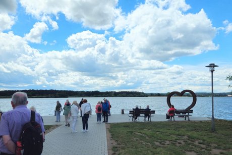 Panoramablick auf den See, im Hintergrund zentral &auml;ltere Menschen, die auf einem Weg in Richtung Seeufer spazieren.