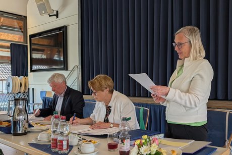 Eine Frau steht an einem Tisch und liest aus einem Dokument vor. Daneben sitzen eine Dame, die Protokoll f&uuml;hrt, sowie ein Mann, der Unterlagen durchgeht.