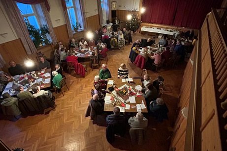 Ein Bild welches von der Empore aufgenommen wurde. Darauf sind 5 Doppeltische mit Personen zu sehen, welche gerade einige Weinachtslieder singen.