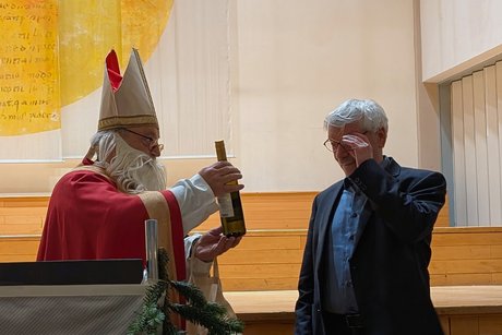 Vorweihnachtlicher Nachmittag Nikolaus überreicht einem Mann im Anzug eine Flasche Wein, der sich mit der linken Hand an die Brille fasst.