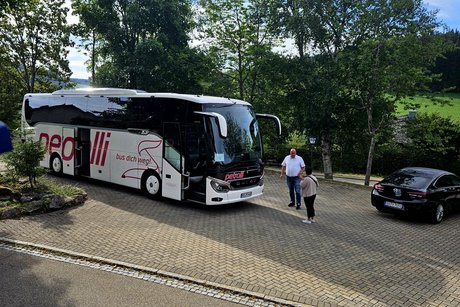 Auf dem gro&szlig;en Parkplatz steht der Reisebus. Der Busfahrer Mike steht davor und spricht mit Michaela.