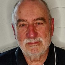 Dieter Sch&uuml;le l&auml;chelnd mit dunklem Poloshirt und leichten wei&szlig;en Strichen, vor einer wei&szlig;en Wand.