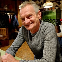 Er sitzt am Wirtshaustisch im grauen pullover und l&auml;chelt.