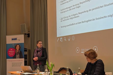 Eine Frau steht vor einer Leinwand mit einer Präsentationsfolie und blickt darauf; neben ihr sitzt eine weitere Frau an einem Tisch und führt Protokoll.