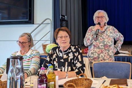 Zwei Frauen sitzen an einem Tisch. Daneben steht eine dritte Frau und hält eine Rede.