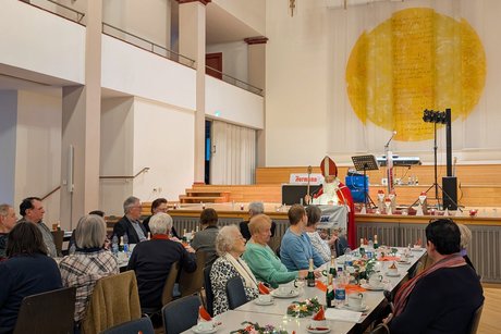 Im Hintergrund steht der Nikolaus an einem Rednerpult. Davor Tischreihen, an denen Gäste sitzen und ihm aufmerksam zuhören.