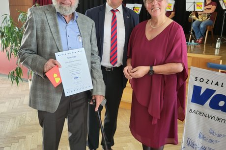 Herr Wolfgang Riekenberg hält eine Urkunde und daneben stehen Reiner Neumeister und Johanna Kern