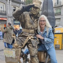 Angela Gutmann auf dem R&uuml;cksitz eines Fahrrades, vorne eine salutierende Lebendstatue im Postdress mit hoher M&uuml;tze in Madrid gem&auml;&szlig; Korbaufschrift des Fahrrades