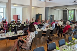 Mehrere Personen sitzen an Tischen in einem großen Saal mit hoher Decke.