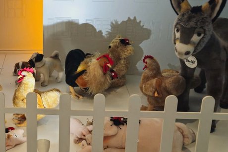 Bauernhof-Stofftiere hinter einem Holzzaun, darunter ein Esel, ein Hahn, ein Schwein mit Ferkeln, eine Henne, ein Mops und ein Teddybär