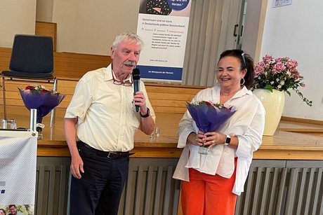 &Auml;lterer Mann mit grauem Schnauzer bedankt sich bei der neben ihm stehenden Frau, die einen Blumenstrau&szlig; in den H&auml;nden h&auml;lt.