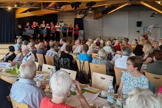 Panorama des Festsaals mit mehreren Tischreihen, an denen viele Leute sitzen. Im Hintergrund die Bühne mit einem Chor.