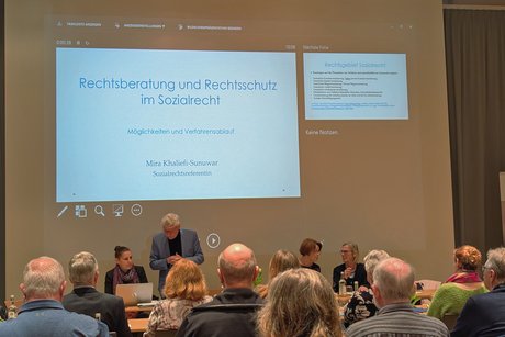 Blick in den Vortragssaal von hinten auf mehrere Tischreihen mit Konferenzteilnehmenden; vorne zeigt eine Leinwand das Thema der aktuellen Präsentation „Rechtsberatung und Rechtsschutz“.
