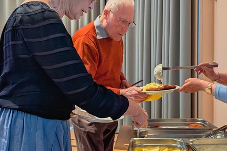 Eine Frau und ein Mann stehen am Buffet; die Frau hält einen Teller, auf den mit einer Suppenkelle Kartoffelsalat serviert wird.