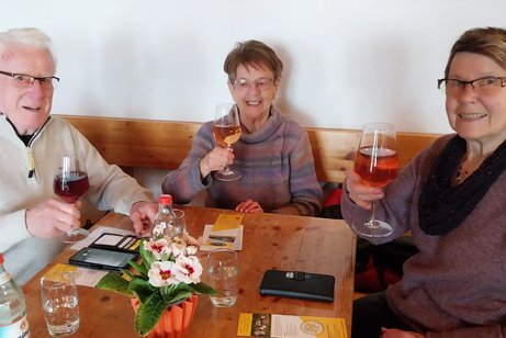 Ein Mann und zwei Frauen sitzen an einem Tisch und prosten mit ihren Weingläsern in Richtung Kamera.