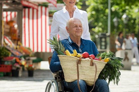 Ein &auml;lteres Paar drau&szlig;en unterwegs, sie schiebt ihn im Rollstuhl. Er hat gef&uuml;llte Einkaufst&uuml;ten auf dem Scho&szlig;.